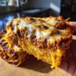 A hand holding a thick slice of baked spaghetti pie, showing layers of pasta, rich meat sauce, and melted mozzarella cheese topping.