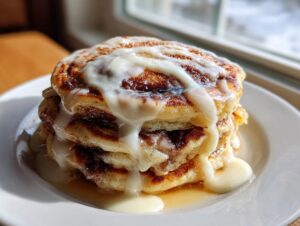 Close-up of a tall stack of fluffy cinnamon roll pancakes drizzled heavily with white cream cheese icing.