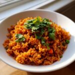 A close-up of fluffy, orange-red spanish rice served in a white bowl, topped with fresh cilantro.