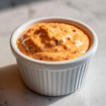 A close-up of creamy, orange-hued smash burger sauce speckled with herbs and red pepper flakes in a white ramekin.