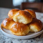A stack of six shiny, golden brown dinner rolls with a visible soft interior on one roll.