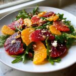 A close-up of a vibrant citrus salad featuring slices of blood orange and navel orange over arugula, topped with feta and pecans.